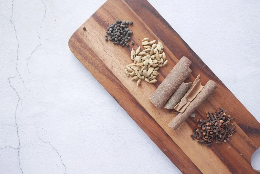 Whole spices on a cutting board: green cardamom pods, cinnamon stick, black peppercorns, cloves, and ginger coins