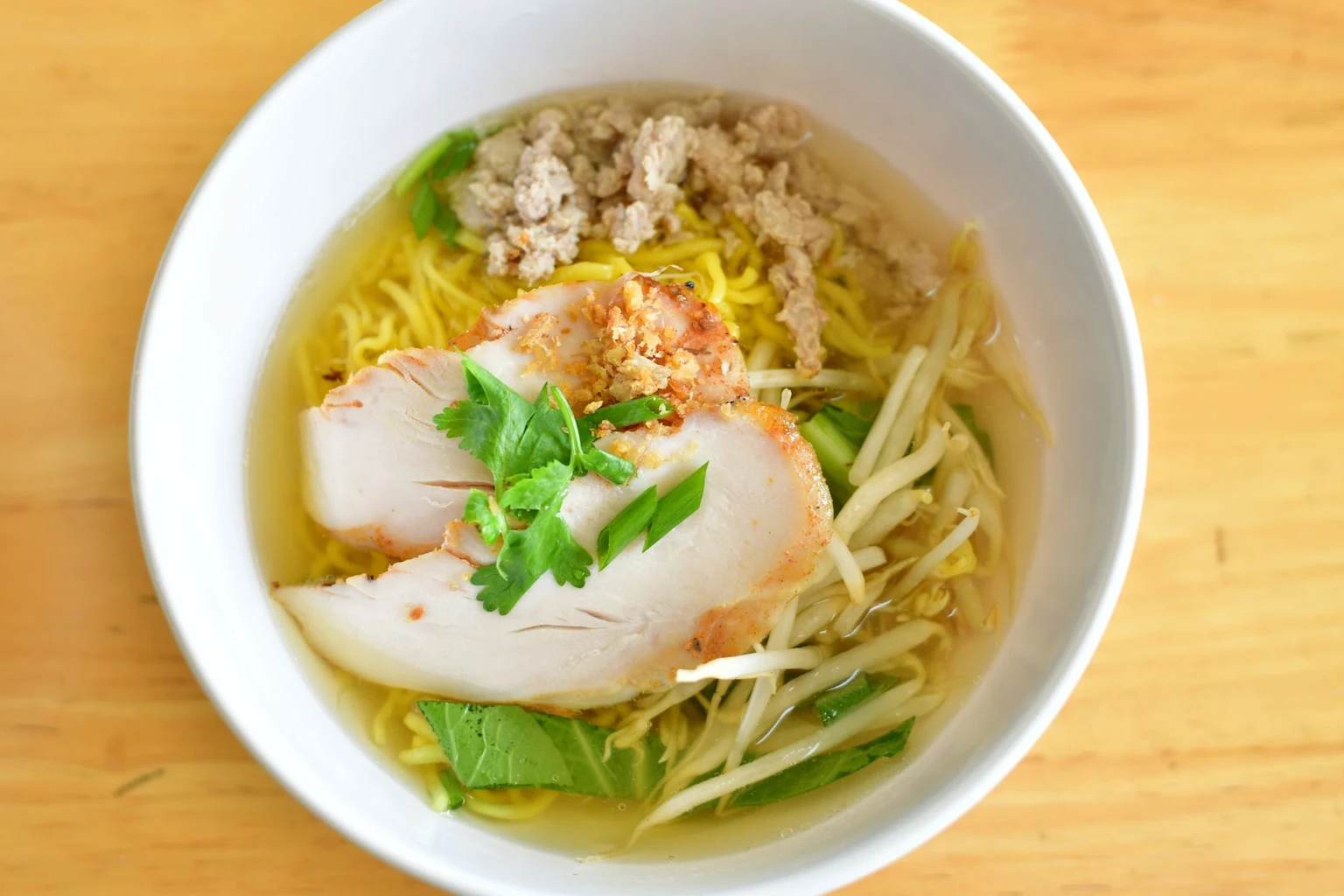 A steaming bowl of vibrant golden-yellow chicken noodle soup with curly egg noodles, shredded chicken, carrots, and cilantro